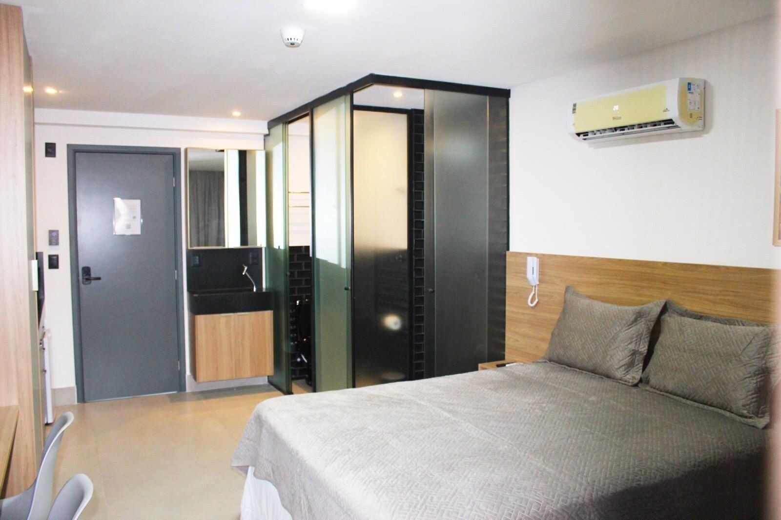 Quarto de hotel com cama, escrivaninha, guarda-roupa e ar-condicionado fixado na parede.