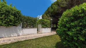 Un sendero pavimentado que conduce a una entrada con portón, rodeado de follaje cuidado y un cielo azul despejado.