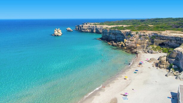Una playa costera con aguas turquesas transparentes, acantilados rocosos y una orilla arenosa con sombrillas y sillas de playa.