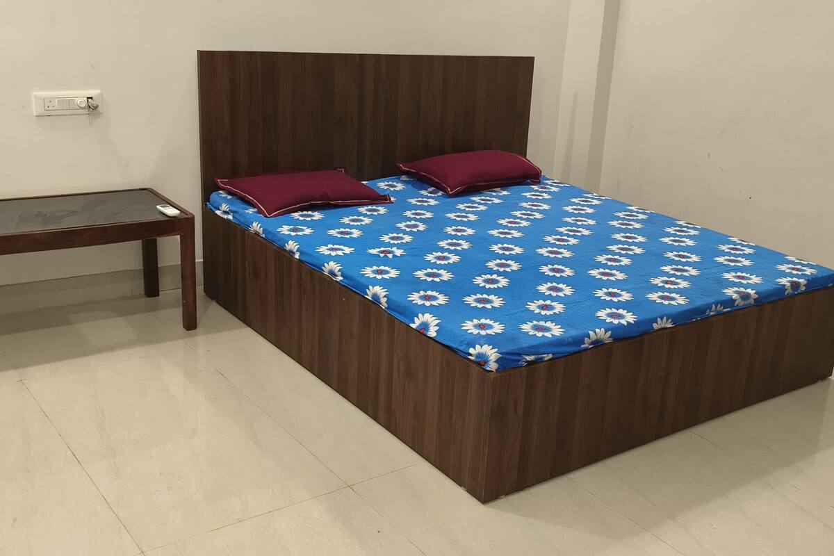 A single bed with a blue floral bedspread and two red pillows, a wooden nightstand, and a wall socket.