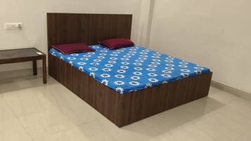 Una cama individual con una colcha azul estampada con flores y dos almohadas rojas, una mesita de noche de madera y un enchufe de pared.