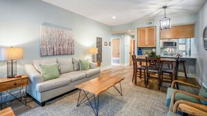 A modern living room with a sofa, coffee table, and dining area. There is a kitchen with wooden cabinets and a dining table. - Camp Warnecke B111- Waters Edge - Stunning Remodel (New Braunfels)