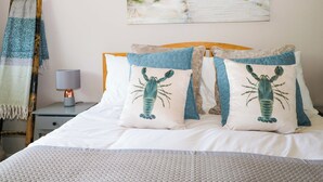 A neatly made bed with two pillows featuring a lobster design, a lamp on a bedside table, and a painting of a beach pathway on the wall. - Pebble Place (Littlehampton)