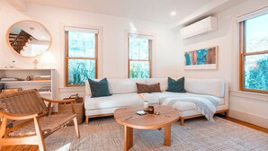 Una sala de estar moderna con un sofá blanco, dos sillas de madera, una mesa de centro redonda y ventanas amplias con vista a follaje.