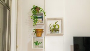 Una esquina de habitación con un armario de madera, una estantería de vidrio con plantas, una mesita con un florero y un televisor.