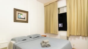 Una cama bien hecha con una colcha azul y dos almohadas, un cuadro en la pared y una ventana con cortinas.