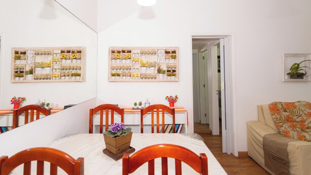 Un comedor con una mesa de madera, sillas y un espejo en la parte superior. Hay plantas en macetas en las paredes y un sofá al fondo.