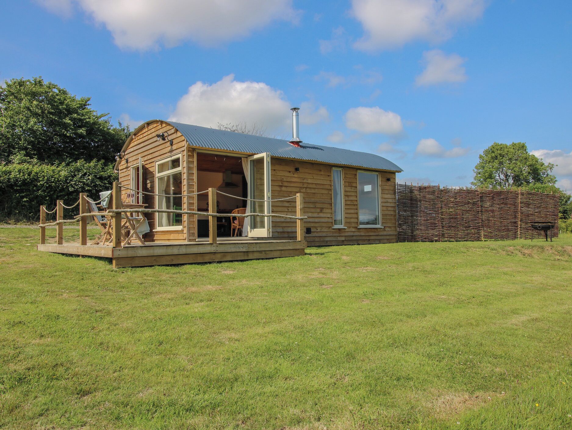 A wooden cabin with a curved roof, a deck with chairs, and a stone wall in the background.