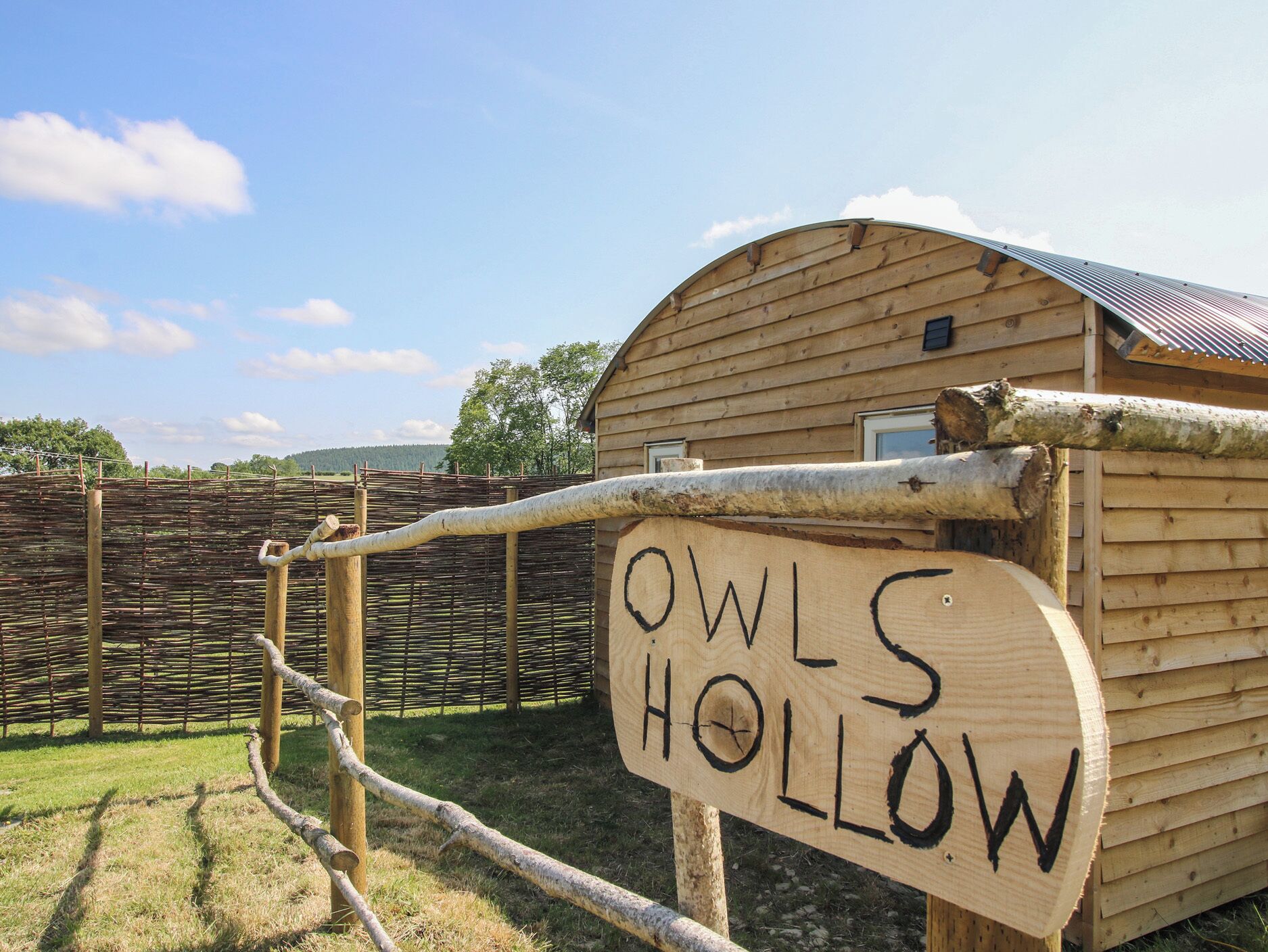 Un letrero de madera con la inscripción 'OWLS HOLLOW', una valla de madera y un edificio con techo inclinado.