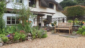 A white house with a thatched roof, surrounded by a garden with various plants and flowers, and a wooden bench in the foreground. - 1 Myrtle Cottages (Newton Abbot)
