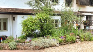 A white building with a thatched roof, surrounded by a garden with various plants and flowers. - 1 Myrtle Cottages (Newton Abbot)