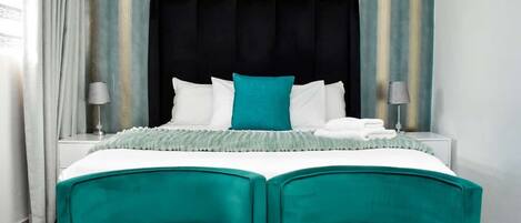 Habitación de hotel con una cama grande, sillas color turquesa, cabecera negra y cortinas a rayas.