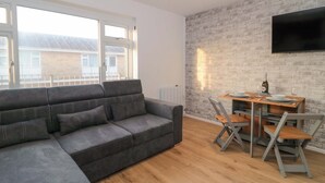 Een moderne woonkamer met een grijze modulaire bank, een houten eettafel met stoelen, een flatscreen televisie bevestigd aan een muur met baksteenmotief, en grote ramen met uitzicht op gebouwen.