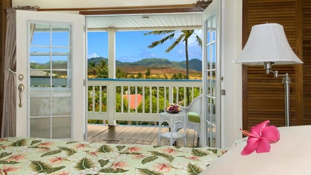 Quarto com cama, uma mesinha e cadeira, varanda com vista para a paisagem e uma luminária.