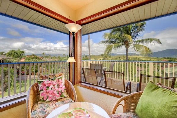 A balcony with wicker chairs, a table, and a view of palm trees and mountains.