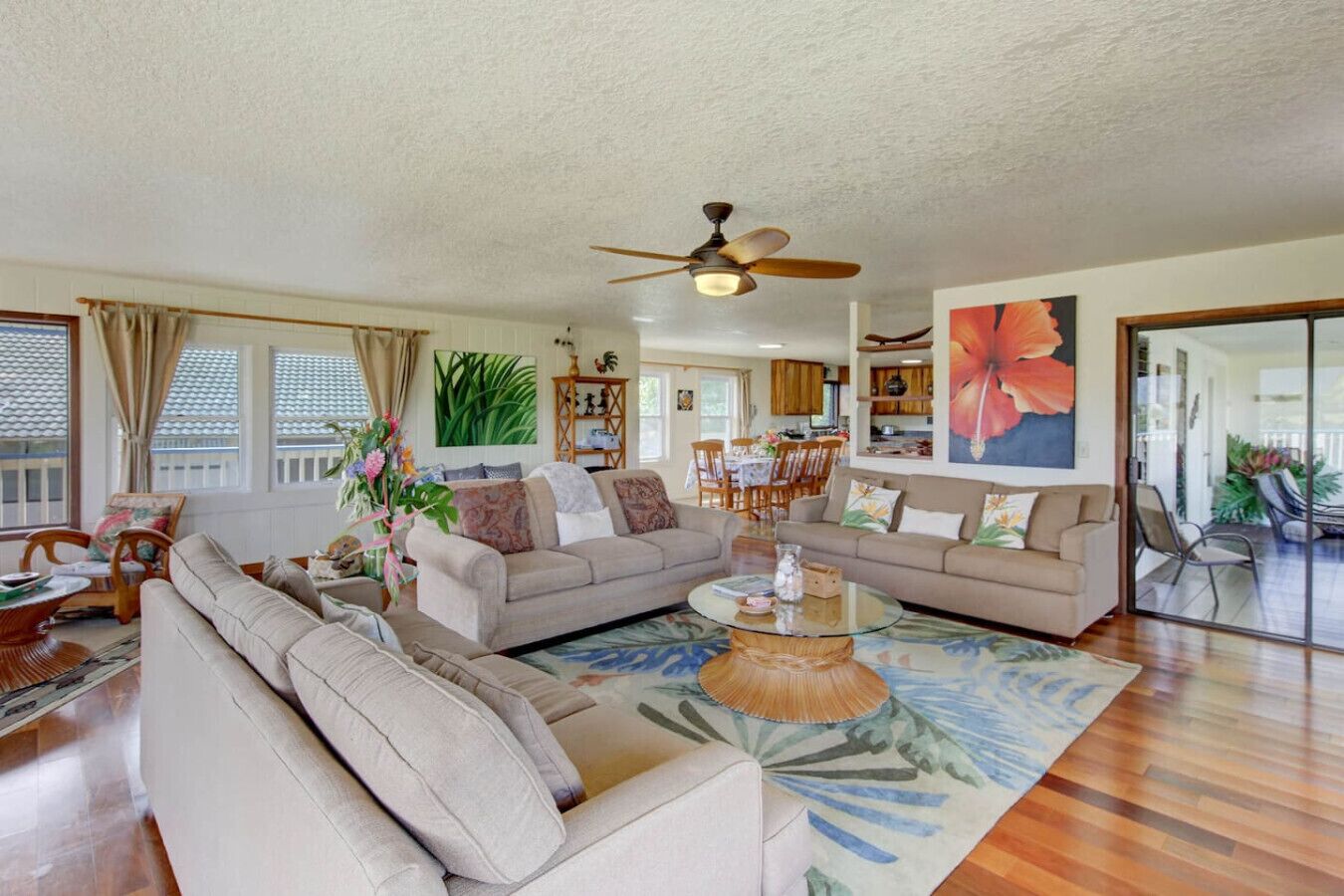 A spacious living room with a large sofa, a coffee table, and a ceiling fan. There is a dining area with chairs and a table in the background.