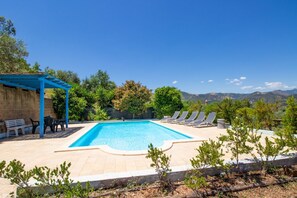 A swimming pool with lounge chairs and a pergola, surrounded by trees and mountains in the background.