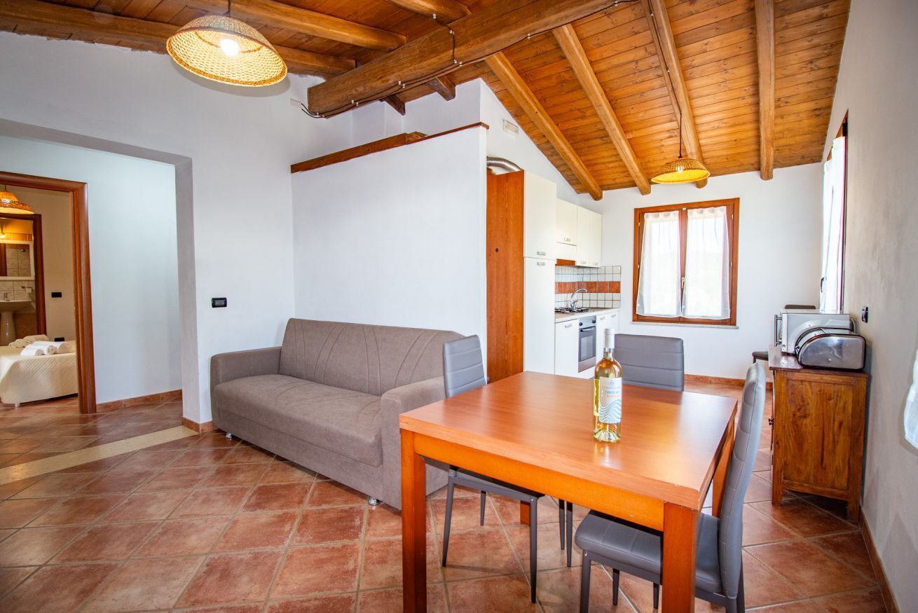 A cozy living area with a wooden ceiling, a sofa, a dining table with chairs, and a kitchen in the background.