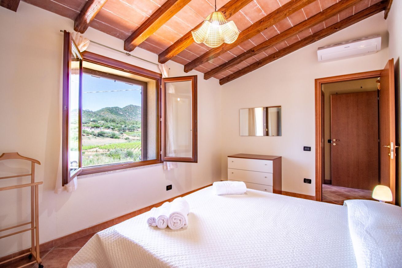 A bedroom with a large bed, a wooden wardrobe, a bedside table, a hanging light fixture, and a window with a view of greenery and hills.