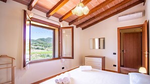 A bedroom with a large bed, a wooden wardrobe, a bedside table, a hanging light fixture, and a window with a view of greenery and hills. - Is Carcuris 1P by All4sardinia  (Cardedu)