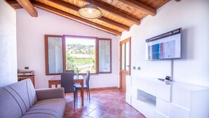 A living room with a sofa, a dining table with chairs, a TV mounted on the wall, and a large window with a view of the countryside. - Is Carcuris 1P by All4sardinia  (Cardedu)