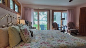 A bedroom with a large bed, a wooden headboard, a window with curtains, a chair, and a table with a lamp.