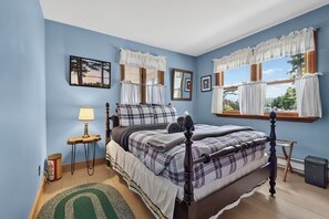 A bedroom with a four-poster bed, a small bedside table with a lamp, a framed picture on the wall, and a window with curtains.
