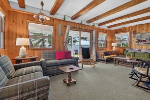Interior acogedor de una cabaña con paredes de madera, un sofá a cuadros, un sillón mecedor y una mesa de comedor.