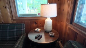 A wooden interior with a plaid couch, a table with a lamp, remote controls, and a small basket.
