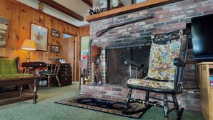 A cozy living room with a brick fireplace, wooden paneling, a green armchair, a wooden table, and a television.