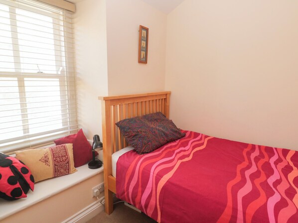Une petite chambre à coucher, chaleureuse et accueillante, avec un lit simple, une fenêtre avec des stores, une table de chevet avec une lampe et un tableau accroché au mur.