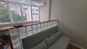 A room with a grey sofa, a window with a view of buildings, and a white air conditioning unit on the wall.