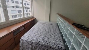 A bedroom with a window, a bed, a bedside table, and an air conditioning unit.