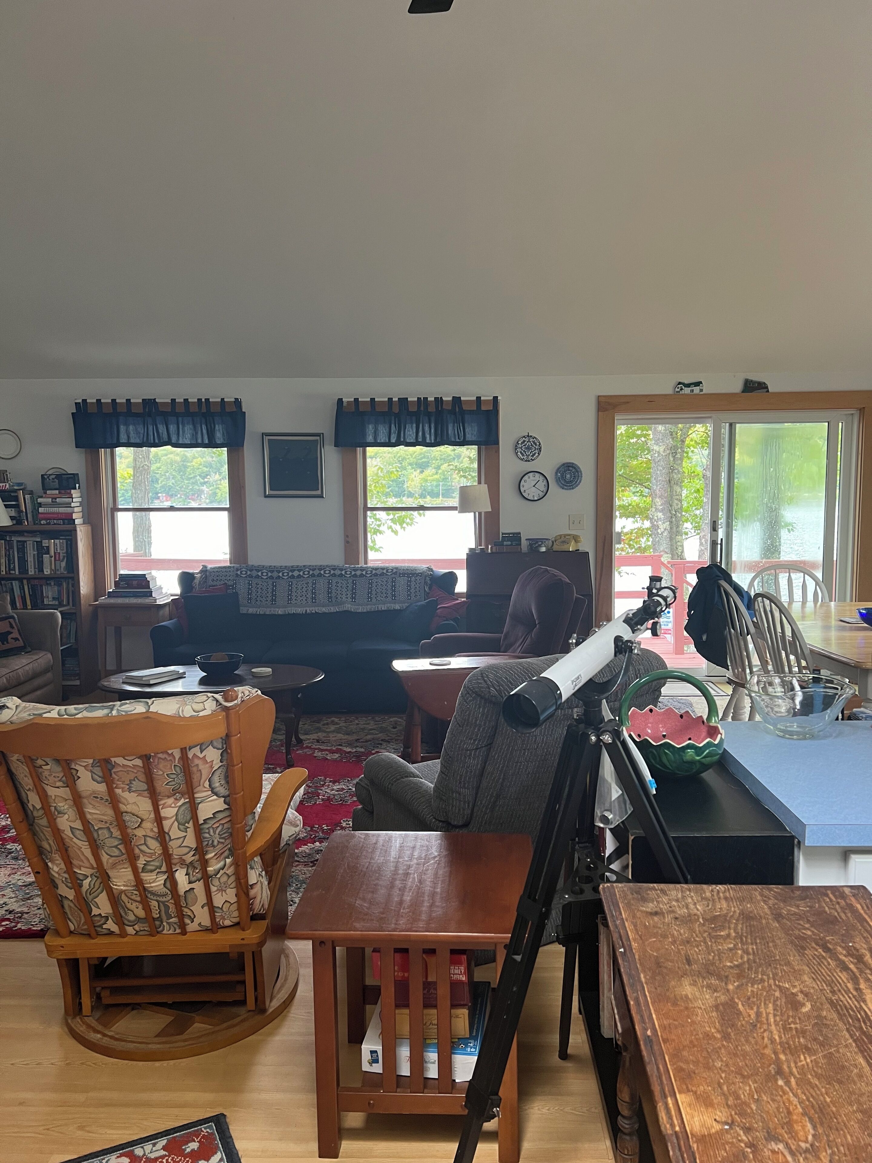 A cozy living room with a wooden rocking chair, a sofa, and a coffee table. There are two windows with curtains, a bookshelf, and a door leading outside.