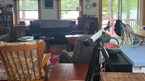 Un salon chaleureux avec un fauteuil à bascule en bois, un canapé et une table basse. Il y a deux fenêtres avec des rideaux, une bibliothèque et une porte qui donne sur l’extérieur.