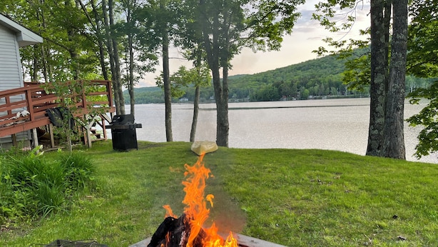 Un foyer avec des flammes, entouré d’herbe et d’arbres, avec un lac et une terrasse en bois en arrière-plan.