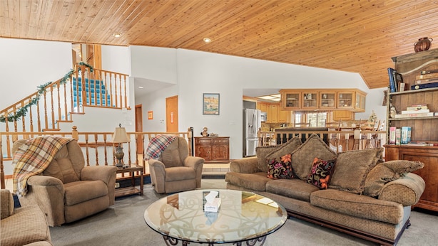 A spacious living room with a wooden ceiling, a round glass coffee table, and two sofas with patterned cushions. A staircase with a garland is visible in the background. - Fox Farm Manor (Big Bear Lake)