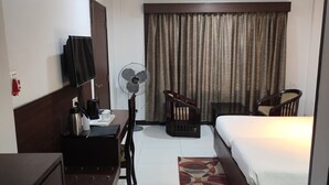A hotel room with a bed, a desk with a chair, a standing fan, a television mounted on the wall, and a window with curtains. - Airport City Hotel (Barakpur)