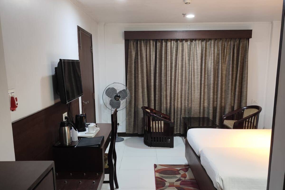 Une chambre d’hôtel comprenant un lit, un bureau avec une chaise, un ventilateur sur pied, un téléviseur fixé au mur et une fenêtre avec des rideaux.