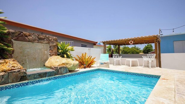 A swimming pool with a tiled edge, surrounded by rocks and plants. There are white chairs and a small table under a wooden pergola. - R&V Combate Beach House, 2nd Floor with Pool (Cabo Rojo)