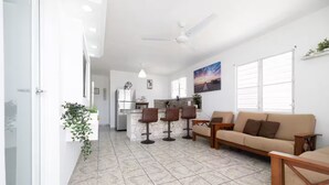 A modern living room with a sofa, two bar stools, and a dining area with a kitchen in the background. - R&V Combate Beach House, 2nd Floor with Pool (Cabo Rojo)