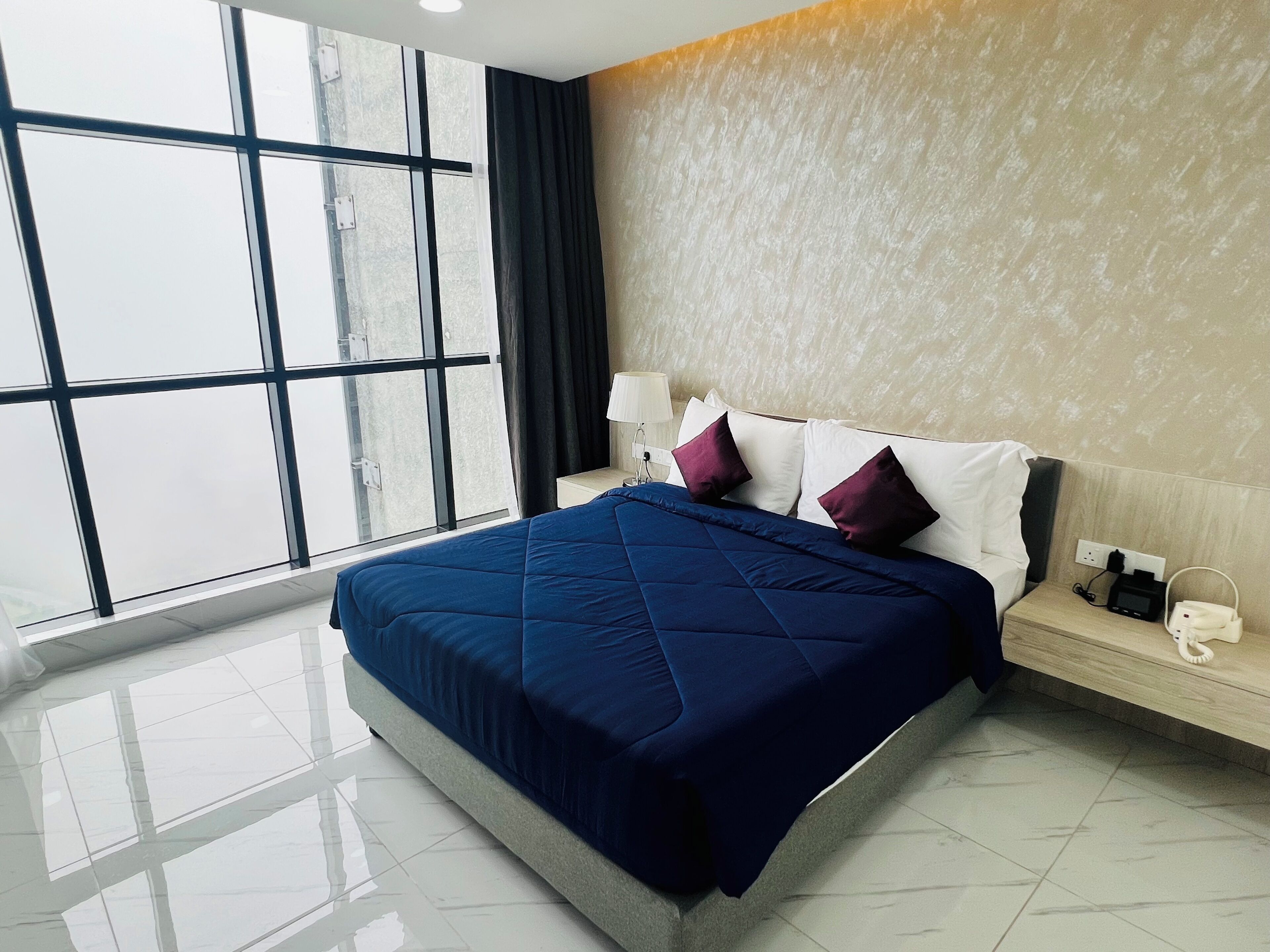 A modern hotel room with a large bed, a nightstand, a lamp, and a view through large windows.