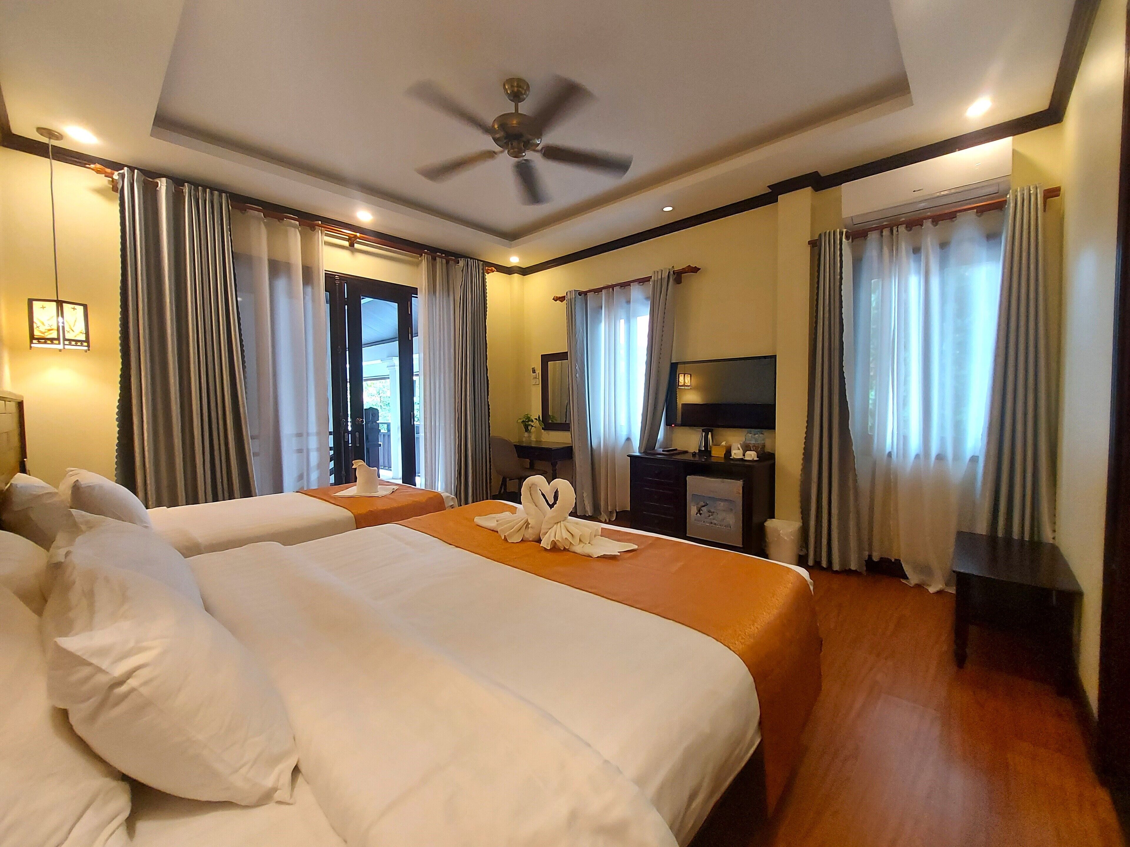 Ein Hotelzimmer mit zwei Betten, einem Deckenventilator, einem Fernseher und einem Schreibtisch mit Stuhl.