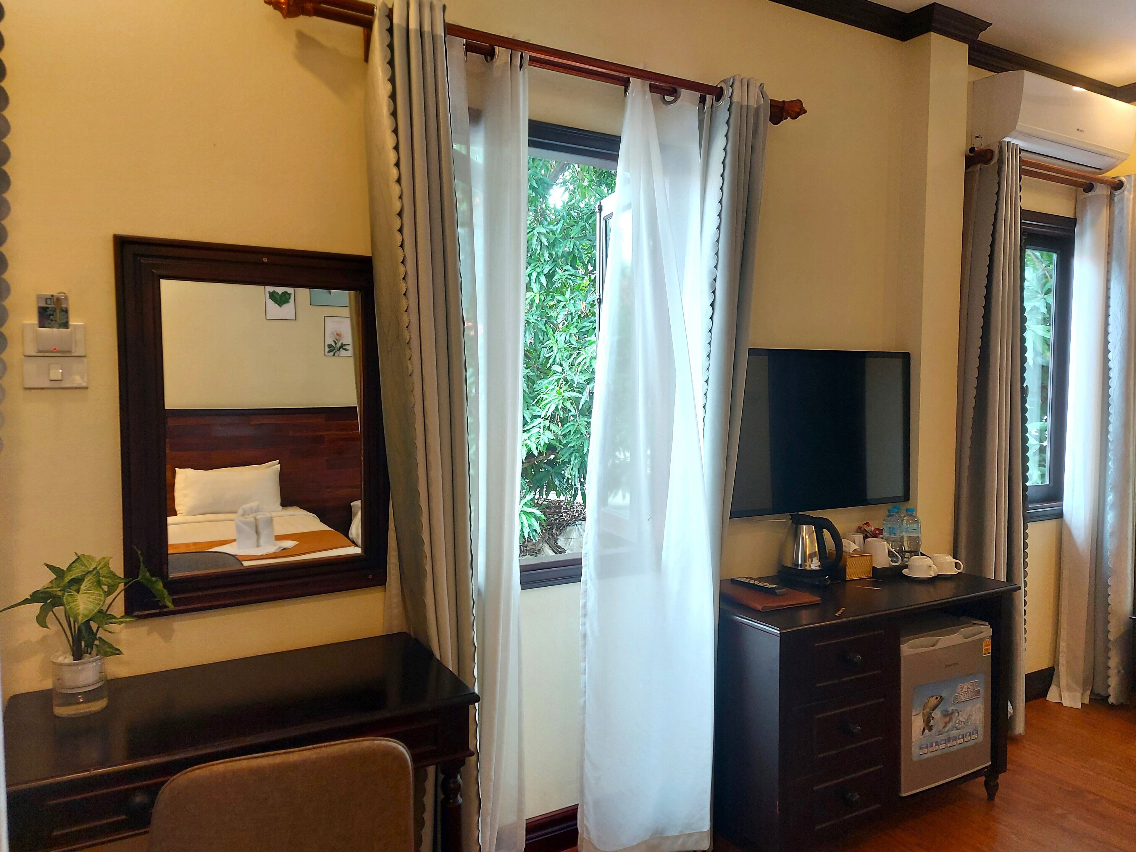 Quarto de hotel com escrivaninha de madeira, televisão, uma pequena planta e janela com cortinas.