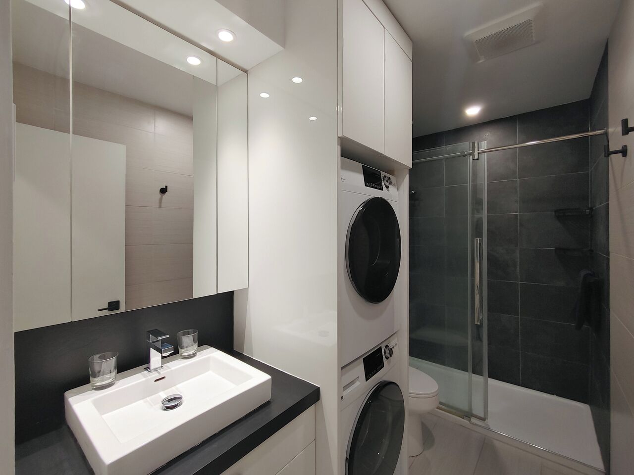 A modern bathroom with a large mirror, a sink, a washing machine, and a glass-enclosed shower.