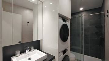 A modern bathroom with a large mirror, a sink, a washing machine, and a glass-enclosed shower.
