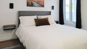 Een netjes opgemaakt bed met een witte dekbedovertrek, een bruin kussen en een grijs hoofdeinde. Daarboven hangt aan de muur een schilderij van een pad door hoog gras.