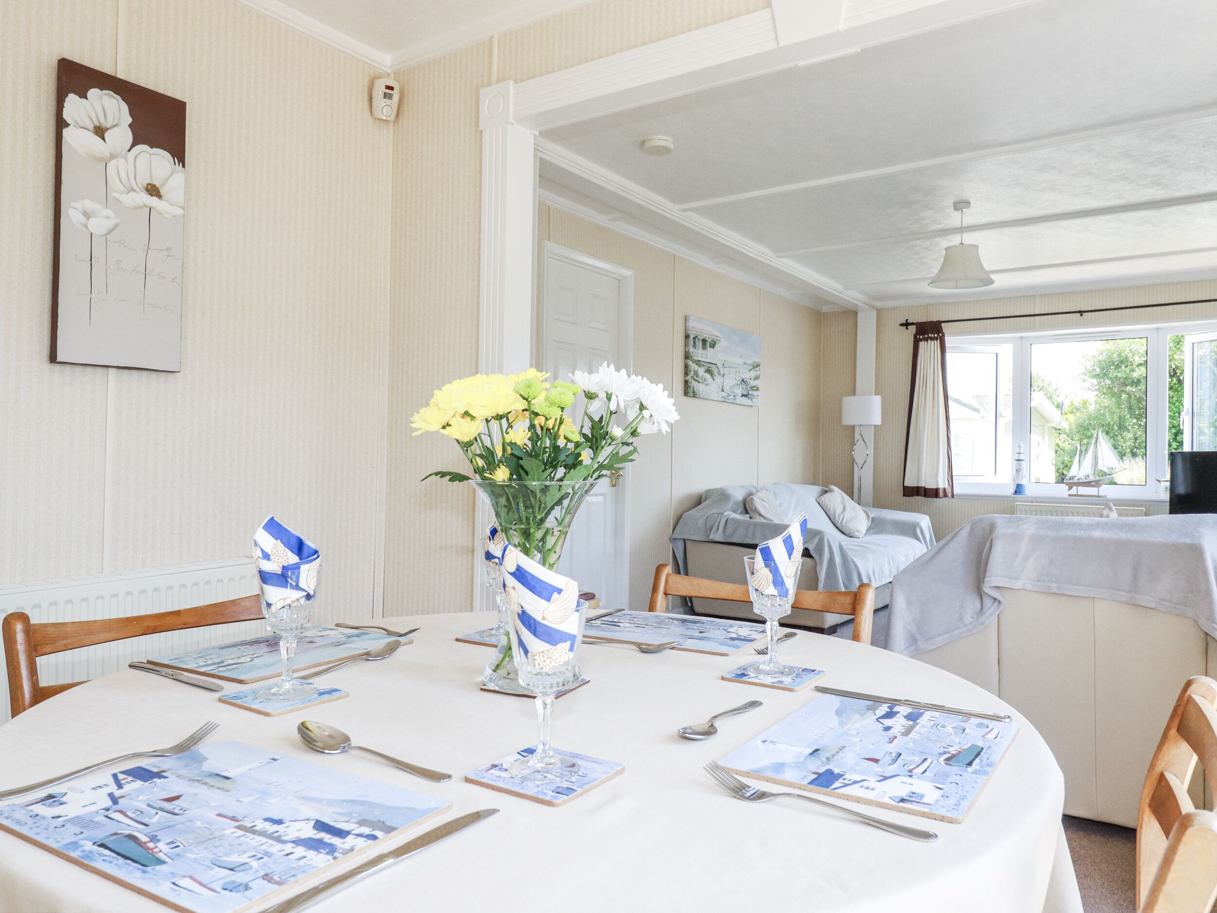 A dining area with a round table set for four, featuring a white tablecloth and blue and white striped napkins. A vase with yellow flowers is placed in the center.