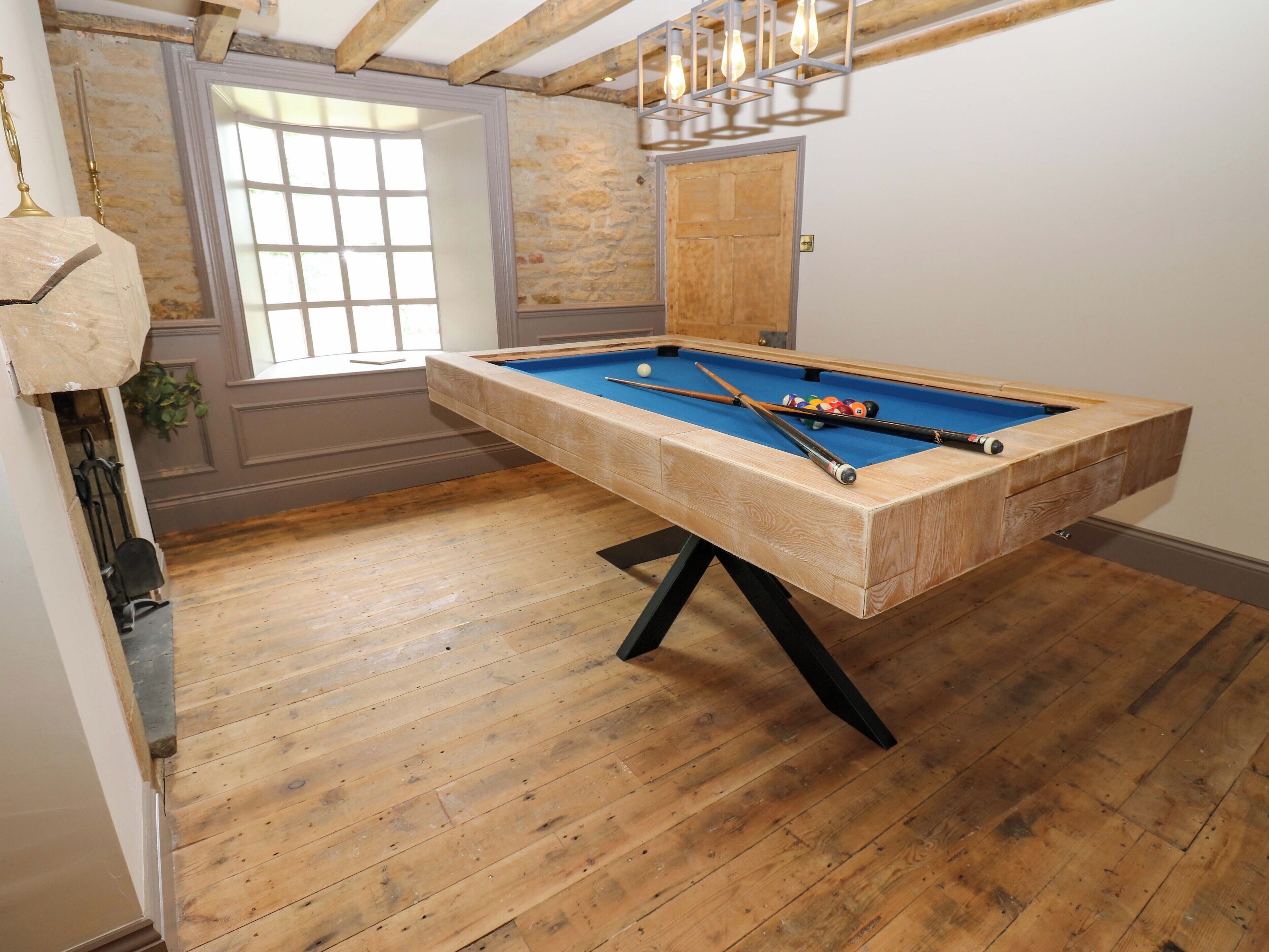 A pool table in a room with wooden flooring, a window, and a stone wall.