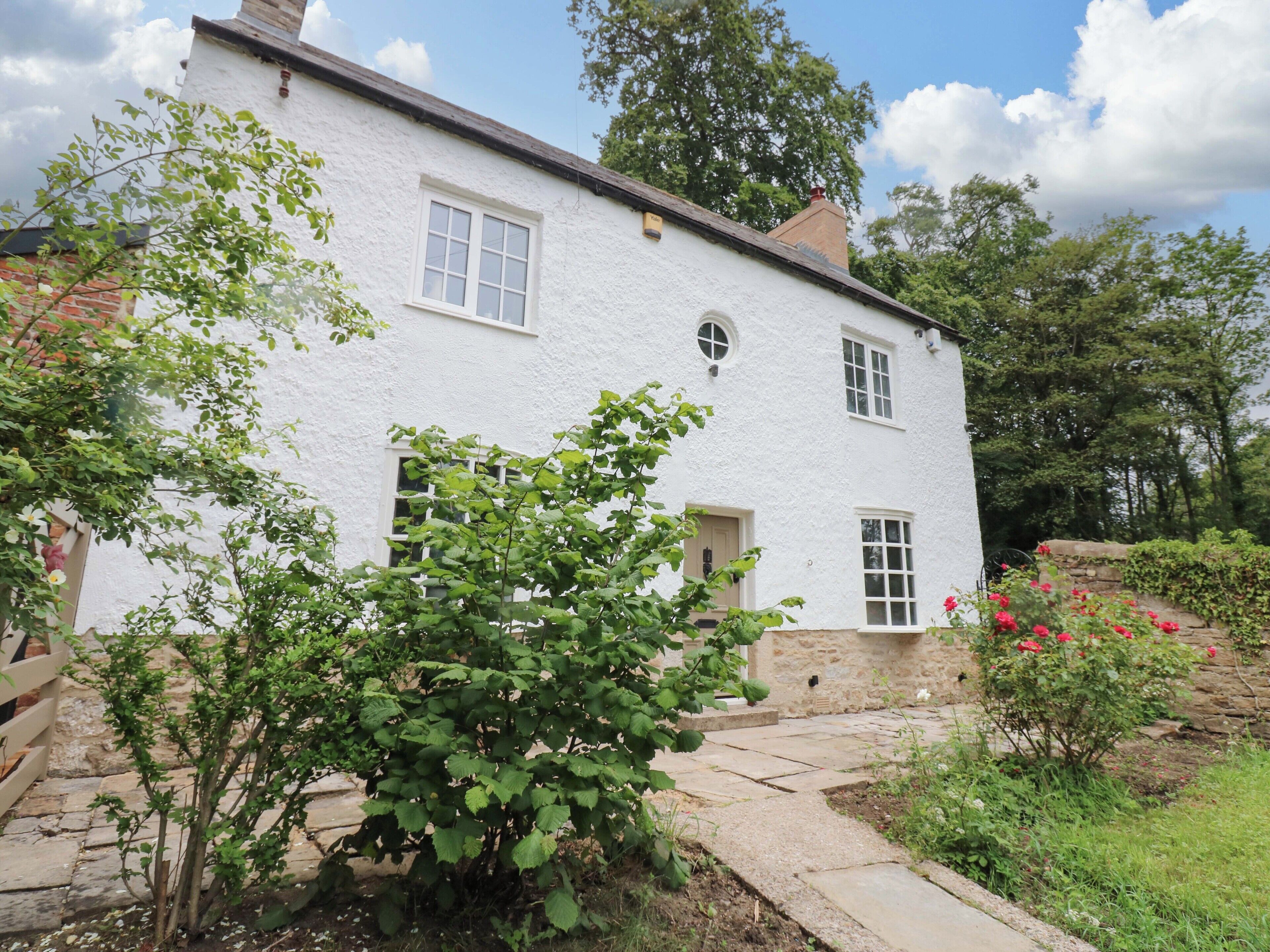 A white two-story house with a stone foundation, a brick chimney, and a small garden with flowering plants.